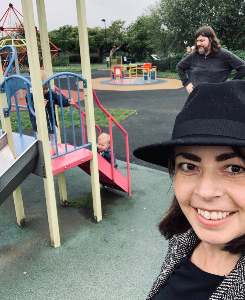 Two young children wearing raincoats play on a colourful playground slide. Woman, front right, taking the selfie, smiling, wears a black fedora. Behind her a man with long hair and beard has hands on waist while looking over the children. It is a rainy day.
